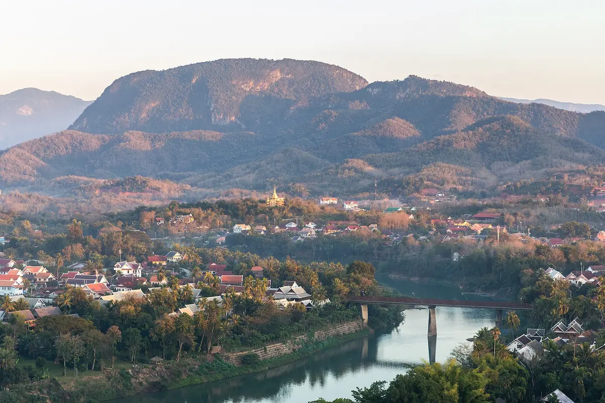 Hory, domy s červenými střechami a most přes řeku Nam Khan v Luang Prabang, pohled z hory Mount Phou Si při západu slunce.