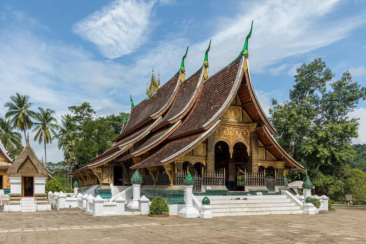 Vícestupňové, obloukovité střechy sim ve Wat Xieng Thong s pozlaceným předním štítem, palmami a menší svatyní vlevo.