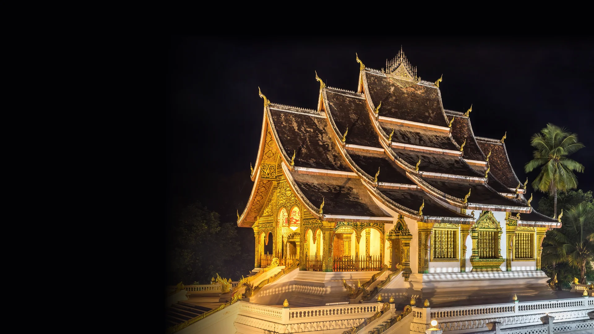 Pozlacené třístupňové střechy chrámu Haw Pha Bang v Luang Prabang, osvětlené proti noční obloze, s palmami vpravo.